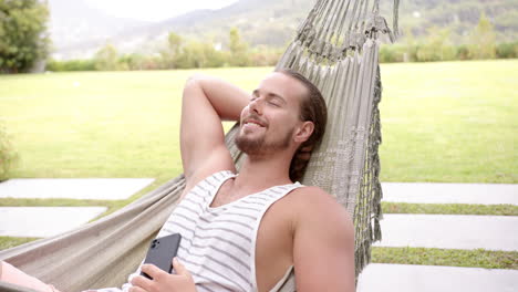 a young caucasian man relaxes in a hammock in the backyard at home, smartphone in hand