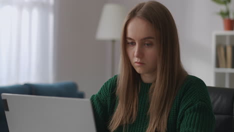 una joven bonita vestida con un suéter verde se está comunicando por chat en línea en redes sociales escribiendo en una computadora portátil retrato femenino interior