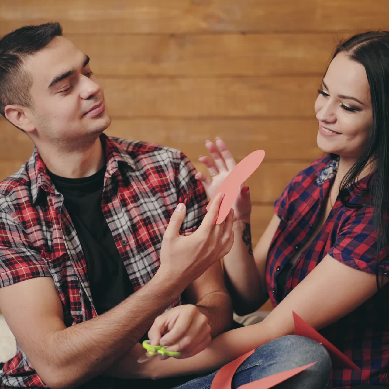 Happy enamored couple are cutting paper hearts from a red cardboard on the bed on the background of a wooden wall in the room. The woman in the plaid shirt is smiling sweetly to man. Preparing for Valentine's Day