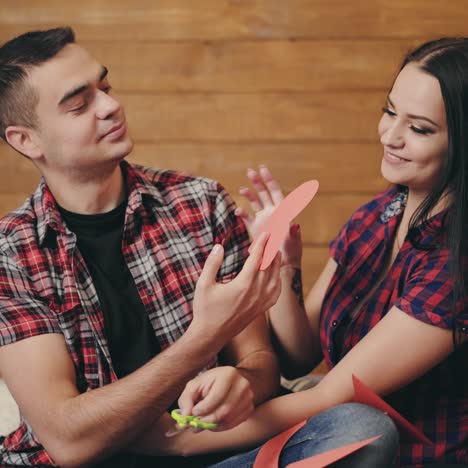 happy enamored couple are cutting paper hearts from a red cardboard on the bed on the background of a wooden wall in the room. the woman in the plaid shirt is smiling sweetly to man. preparing for valentine's day