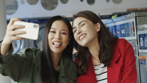 And-Young-Beautiful-Girls-Smiling-And-Posing-To-Smartphone-Camera-While-Taking-Selfie-Photo-In-Laundry-Service