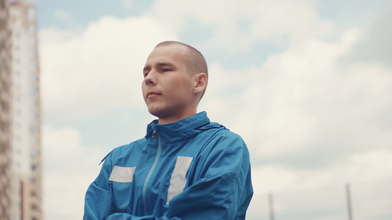 young man in blue rain jacket by urban skyline, profile and threequarter shots, cloudy horizon, city towers framing subject, contemplative expression, quiet anticipation before match, cinematic