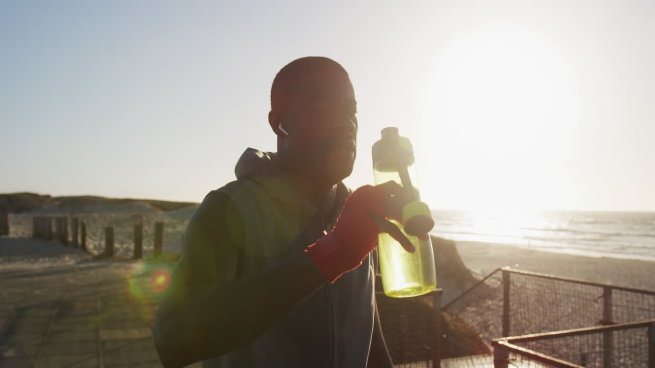 hombre afroamericano vertiendo agua en su cabeza, tomando un descanso en el ejercicio al aire libre junto al mar