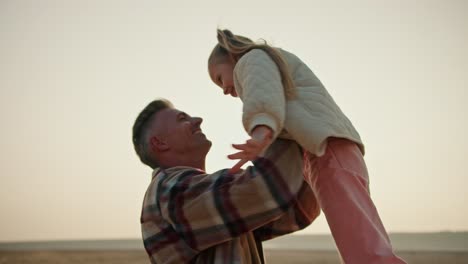 A-happy-middle-aged-brunette-man-with-gray-hair-in-a-plaid-shirt-lifts-his-little-daughter-a-blonde-girl-in-a-white-jacket-and-spins-with-her-during-his-vacation-on-a-hike-on-a-deserted-seashore-in-a-summer-evening