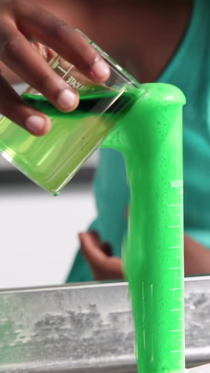 Alumno con gafas vertiendo el contenido de un vaso de precipitados turquesa inclinado en una probeta de laboratorio, demostración de desbordamiento de espuma, con espacio para texto.