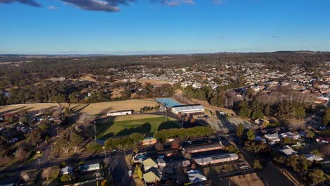drone camera smoothly pans over a sunlit small town, showcasing residential areas, sports fields, and surrounding wooded hills under clear blue skies