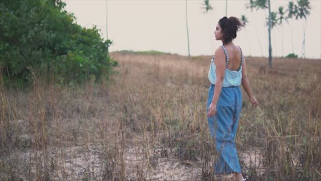 Rearview-of-a-female-walking-along-meadow-among-dry-grass,-hot-summer-day