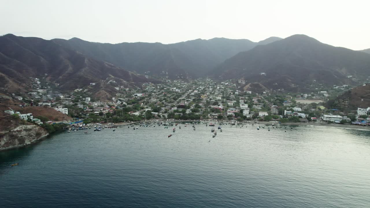 A cinematic aerial panoramic view panning across the picturesque Taganga bay, showcasing the vibrant fishing village, the many colorful local boats, and the arid foothills of the Sierra Nevada