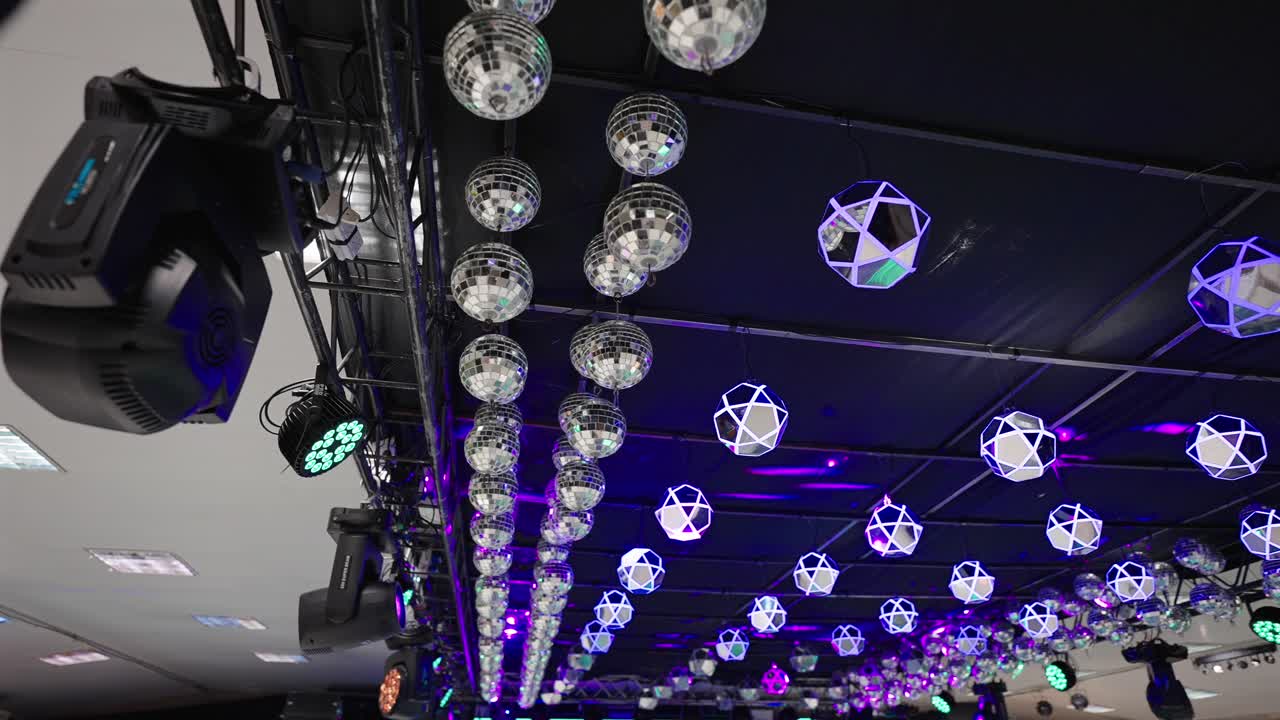 Upward reveal shot showing ceiling filled with mirrored balls and geometric light fixtures, creating reflective light patterns and vibrant atmosphere in an indoor event space