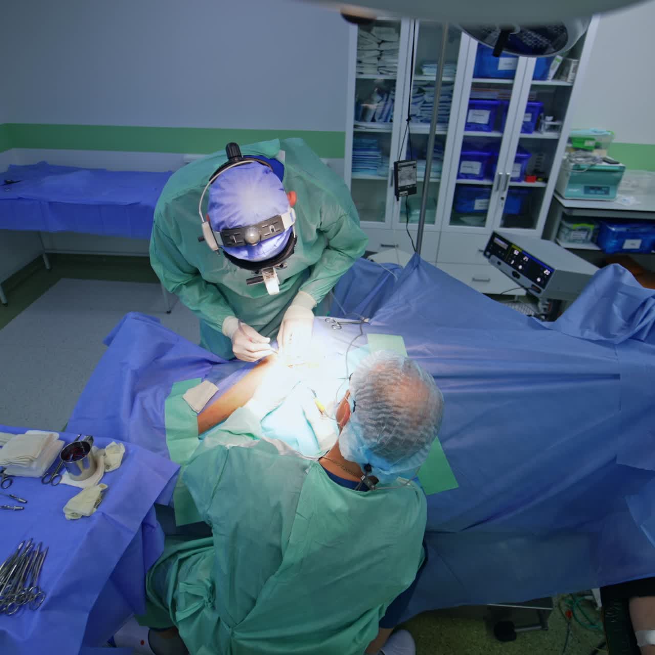 Doctors operate the well-highlighted area on a patient's body. Assisting nurse stands nearby watching intently the surgery. Top view