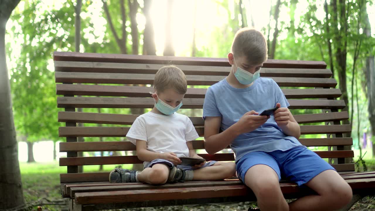 의료 마스크를 입은 소년들이 공원에서 스마트폰으로 놀고 있다.