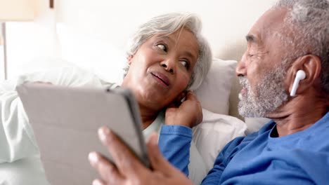 Glückliches-älteres-Gemischtrassiges-Paar,-Das-Auf-Dem-Bett-Liegt,-Tablet-Und-Kopfhörer-Benutzt,-Unverändert,-In-Zeitlupe