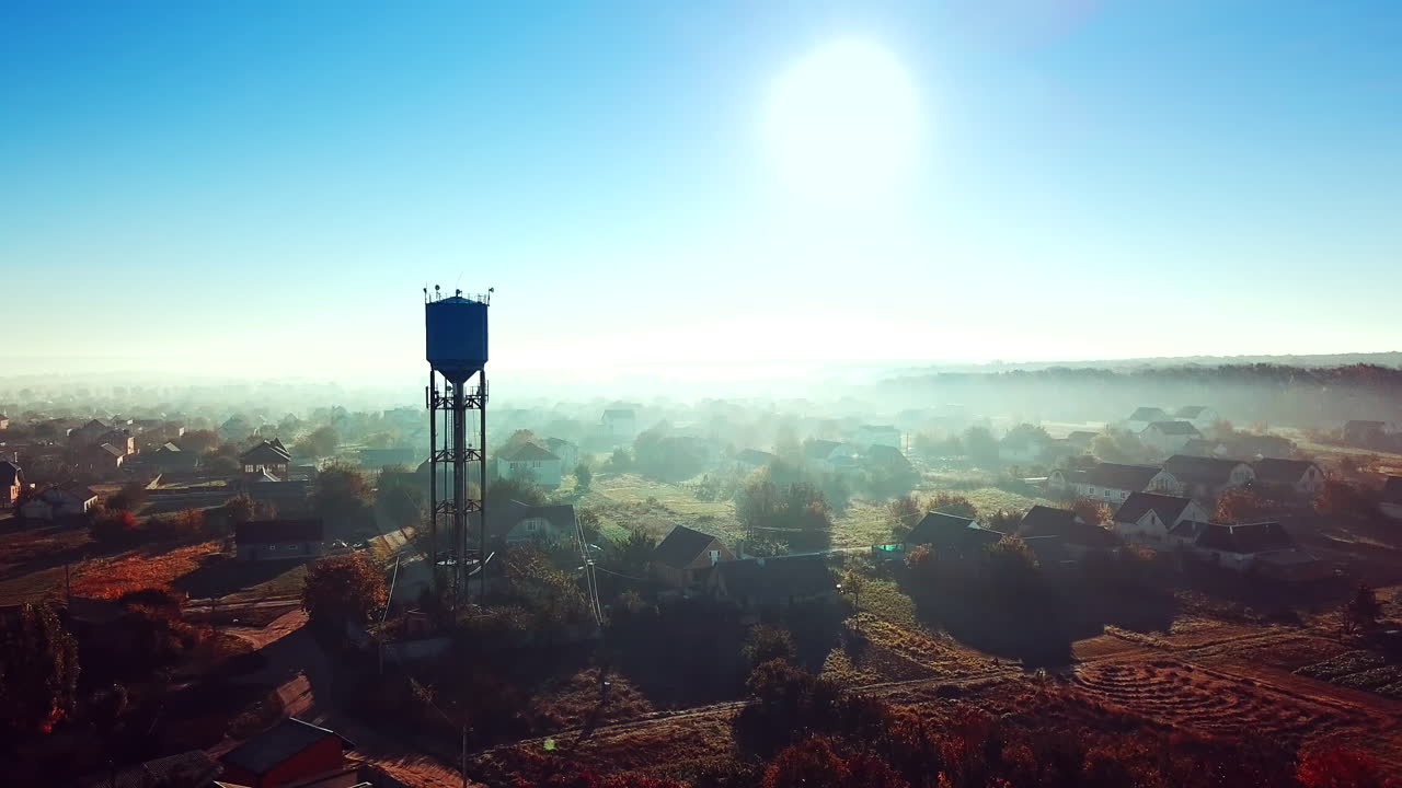 Birds-eye view of a beautiful morning landscape of a rural place. Flying over the small houses with gardens and high construction at sunrise. Aerial view. Camera moves forward. Slow motion.