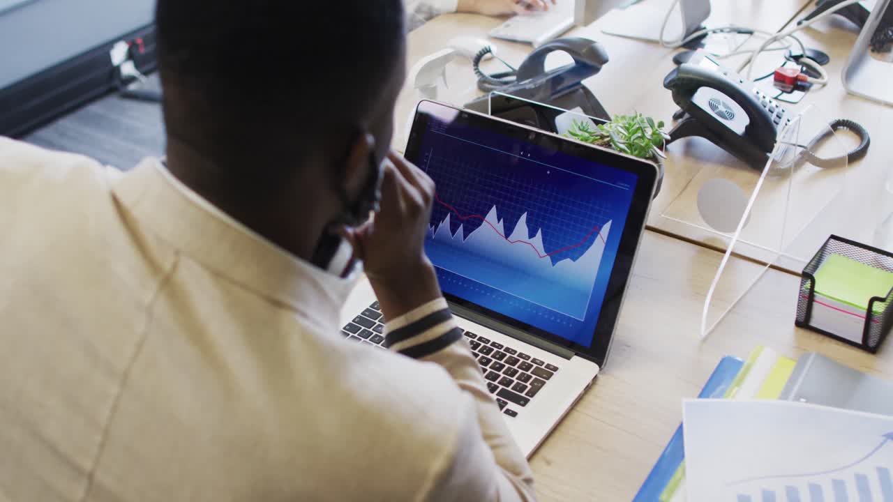 African american man wearing face mask using laptop while sitting on his desk at modern office