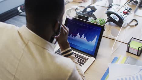 afrikanisch-amerikanischer mann trägt eine gesichtsmaske und sitzt auf seinem schreibtisch in einem modernen büro mit einem laptop