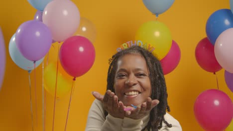 Studio-Portrait-Of-Woman-Wearing-Birthday-Headband-Celebrating-Blowing-Paper-Party-Confetti-To-Camera