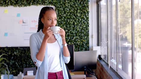 African-american-casual-businesswoman-drinking-coffee-and-looking-out-window-in-office,-slow-motion