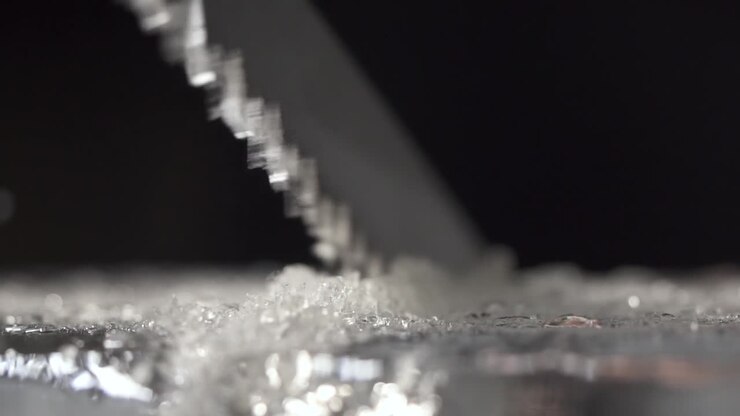 A bartender is cutting the ice block to smaller pieces for cocktail service by using a long toothed knife.