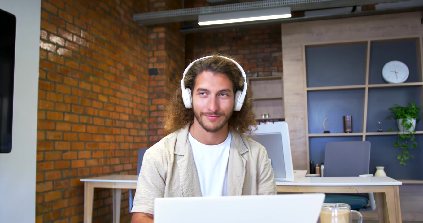 Homem sentado na mesa do escritório, digitando no laptop, atendendo a uma chamada via fone de ouvido, sorrindo com uma caneca.