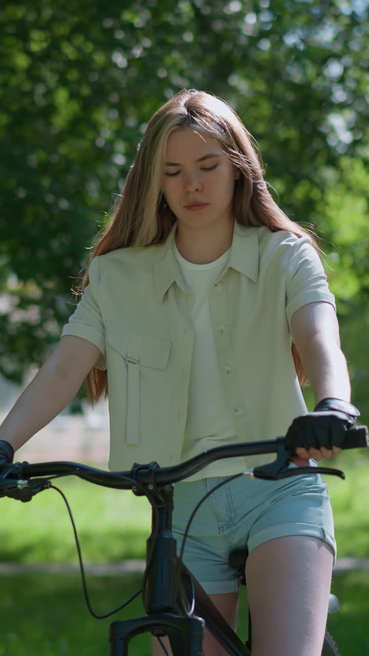 자전거 장갑을 입은 젊은 여성, 야외에서 자전거에 앉아, 그녀의 손에 집중적으로보고, 배경에는 울창한 초록색, 건물 및 나무가 있습니다.