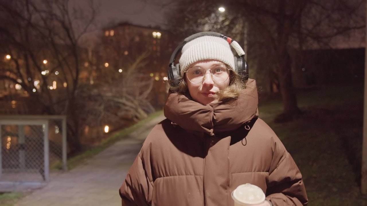 Asian student nighttime park holding coffee, wearing headphones and beanie, glasses fogging slightly, padded coat zipped high, soft streetlight bokeh and pond reflection, slow candid stroll, cinematic