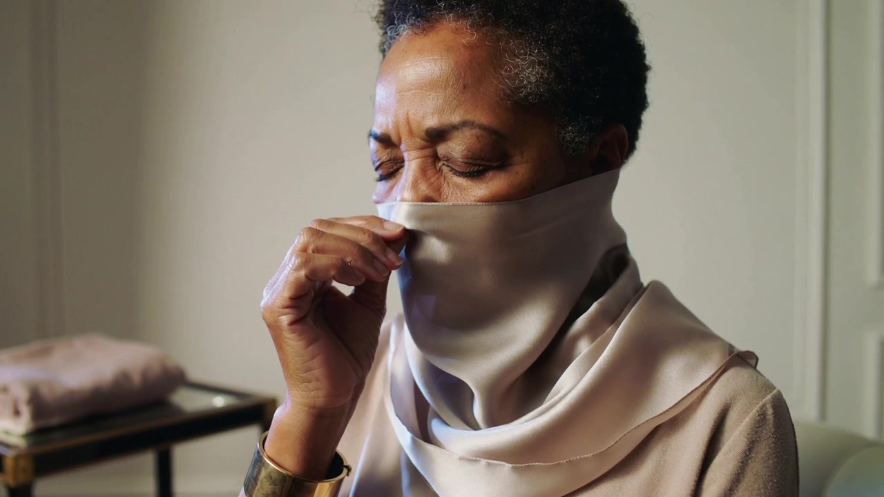 Une femme âgée tient un foulard en soie sur son nez à la maison, montrant un bracelet manchette métallique.