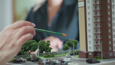 close up hand view of someone holding pencil explaining project to client using detailed architectural building miniature with trees and parked cars in foreground within office setting