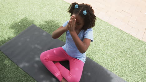 biracial girl meditates on a yoga mat outdoors in school, hands pressed together