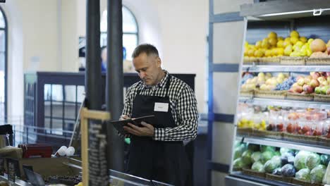 A-middle-aged-grocery-store-worker-continues-inventory-in-the-fruit-section.-A-man-with-a-notebook-and-a-pencil-in-his-hands-is-exposed-to-the-presence-of-products