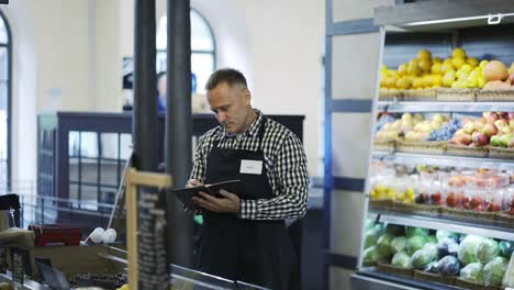 a middle-aged grocery store worker continues inventory in the fruit section. a man with a notebook and a pencil in his hands is exposed to the presence of products