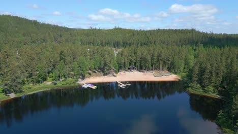 Drone-descends-on-calm-lake-reflecting-pine-tree-forest-and-Sandy-banks-of-resort