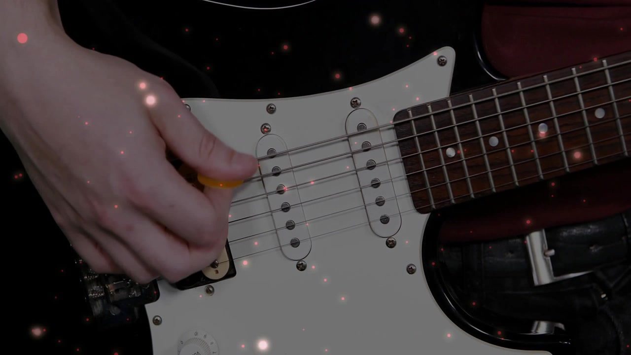 Musicista che pizzica la chitarra elettrica con la mano destra in uno studio casalingo, indossando un top nero, con un plettro giallo.