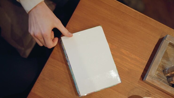 Close up of person hand placing and adjusting objects on wooden table. white book is positioned alongside other items with soft lighting highlighting texture of both hands and materials