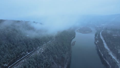 Aerial-footage-of-a-snowy,-scenic-byway,-winding-mountain-valley-road-during-a-snowstorm-with-pine-trees,-a-river,-mountain-highway,-rocky-cliffs,-and-forests-during-winter-on-a-cold,-blue-day