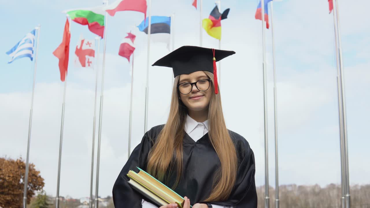 una graduada universitaria de pie en el fondo de las banderas del mundo con libros en las manos. vista del espacio abierto. banderas nacionales en el cielo azul