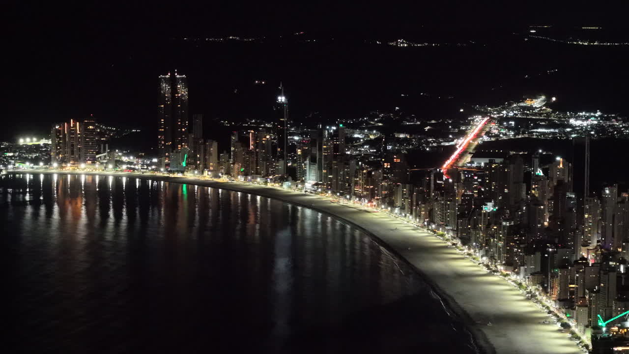 A spectacular aerial shot shows the brightly illuminated skyline of Balneário Camboriú, Brazil at night, with city lights reflecting on the water along the iconic crescent-shaped beach