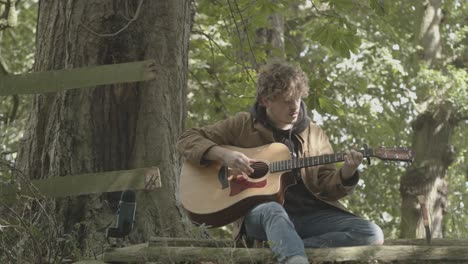 young man plays acoustic guitar and sings sitting under shady tree