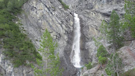 aerial drone footage slowly flying up above a tree canopy to reveal a dramatic waterfall as it cascades over the edge of a sheer mountain cliff into a plunge pool in switzerland