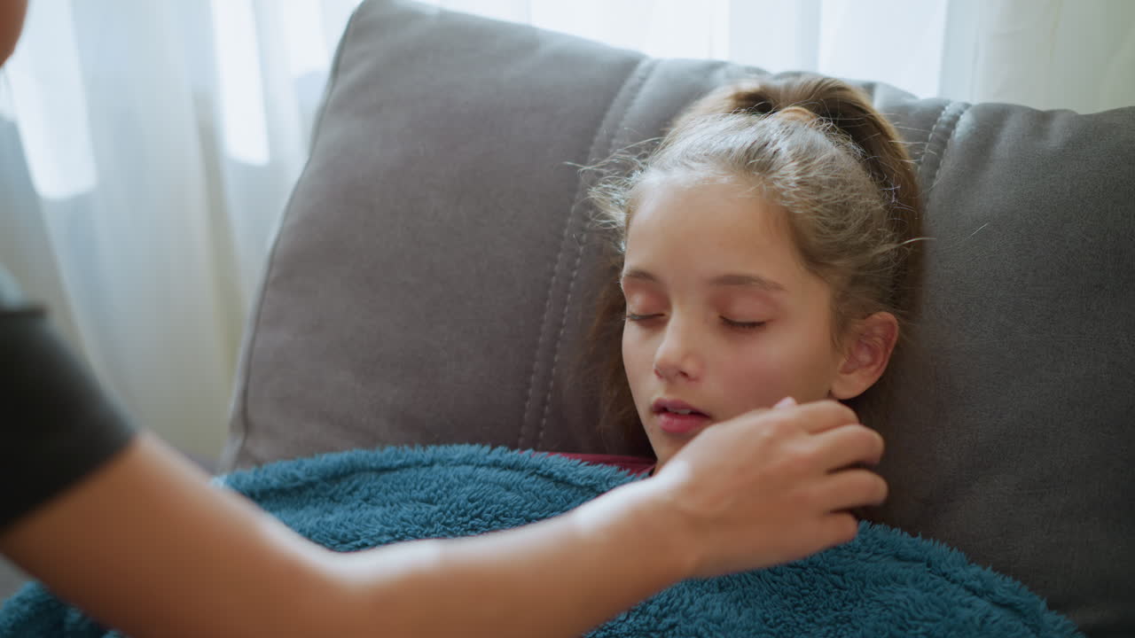 Close up of boy caring for sick sister lying on the couch, gently covering her with a blanket as she opens her eyes and he checks her temperature, showing a nurturing moment