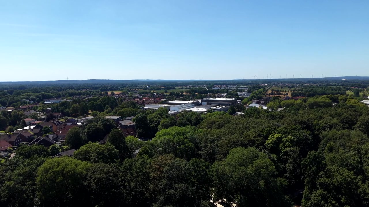 Tiheä metsän latvusto levittäytyy pikkukaupungin kattojen ja teollisuusrakennusten eteen Yhdysvalloissa, kaukaiset tuulivoimalat kohoavat sumuisen puurajan yläpuolelle kirkkaan kesätaivaan alla. Laaja ilmakuva dronella eteenpäin.
