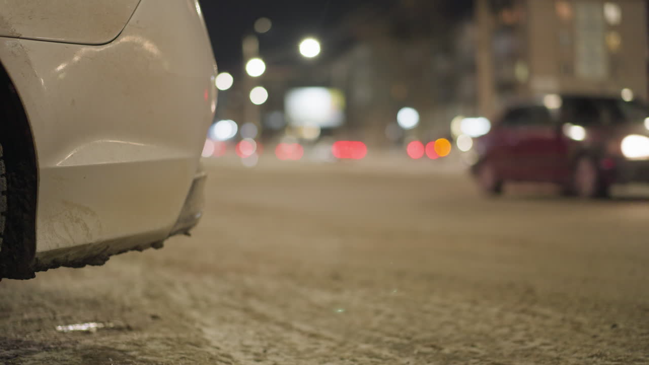 지나가는 자동차와 함께 밤에 고속도로의 가까운 시야, 백색 차의 퍼의 하단 부분에 초점을 맞추고, 배경에는 흐릿한 도시 불빛과 교통이 있습니다.