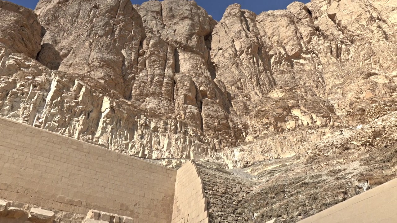 Pengambilan gambar lanskap sudut rendah yang menampilkan tebing kapur Deir el-Bahari yang masif dan curam menjulang di atas dinding penahan batu dan struktur bertingkat Kuil Makam Hatshepsut di Luxor