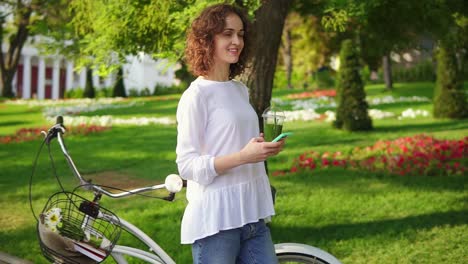 Mujer-Sonriente-Feliz-Enviando-Mensajes-Usando-Su-Teléfono-Inteligente-Parada-En-El-Parque-De-La-Ciudad-Cerca-De-Su-Bicicleta-De-La-Ciudad-Con-Flores-En-Su-Canasta