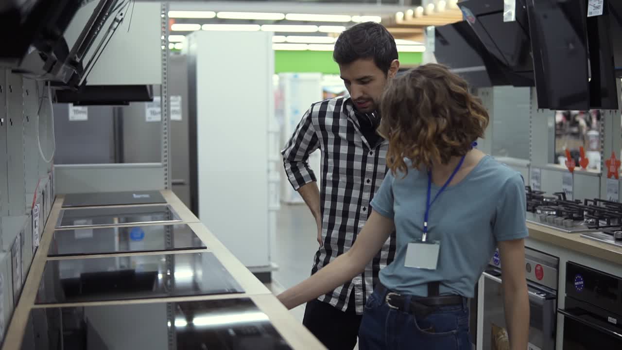 hombre de pelo oscuro, sonriente con camisa a cuadros con auriculares eligiendo una nueva cocina moderna en la tienda. él está planeando comprar muebles y electrodomésticos, comprobando y asesorando con una consultora femenina sobre el horno