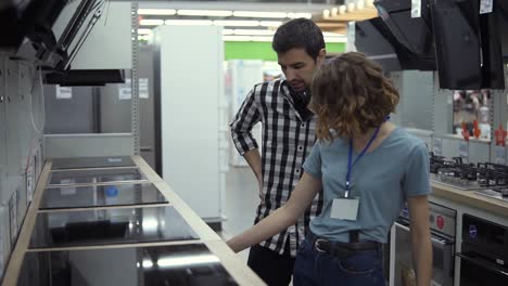 dark haired, smiling man in plaid shirt with headphones choosing a new modern cook top in store. he is planning to buy furniture and home appliances, checking and advising with female consultant about oven