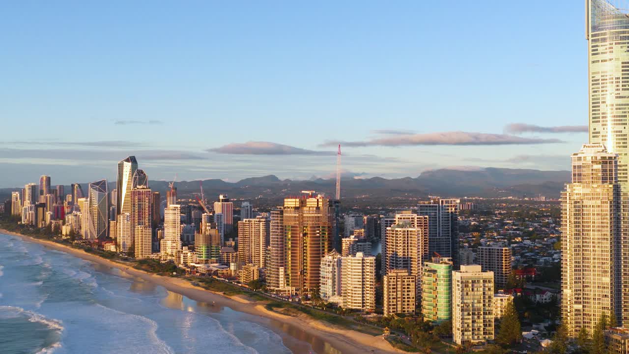 Aerial footage captures Gold Coast's skyline and coastline during sunset, highlighting tall buildings and the ocean under warm lighting