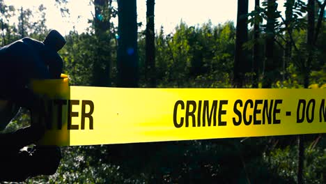 close up view of a police detective or investigator unravel or unrolling and pulling a yellow colored crime scene tape in front of a wooded area or a forest during a sunny day, tape is backlit by sun