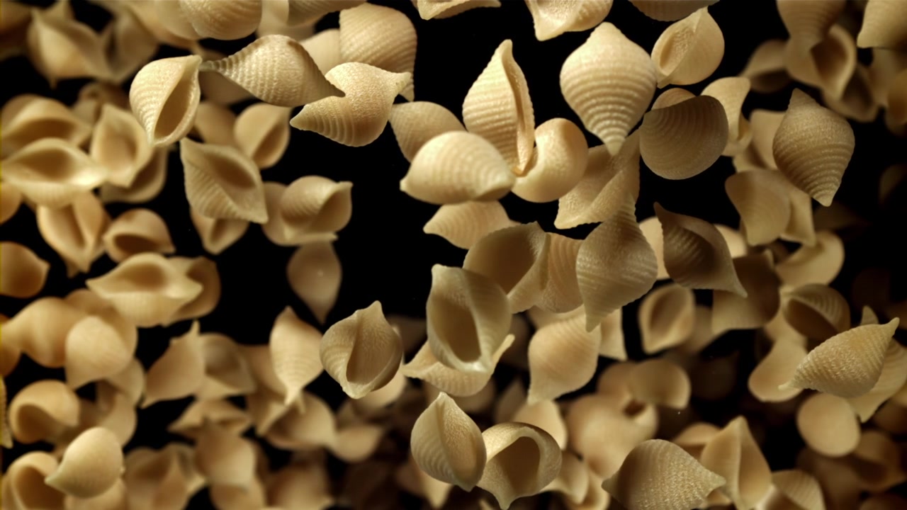 Pasta conchiglie secca che cade al rallentatore su sfondo nero.