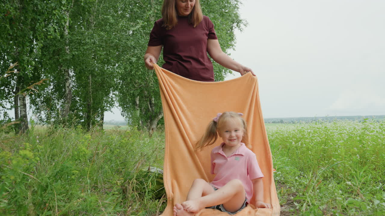 white mother covering daughter with blanket, lakeside meadow peekaboo moment, mother tucks towel around child, gentle hands, giggling toddler, warm summer air, candids of bonding, nurturing gesture