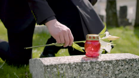 검은 양복을 입은 알아볼 수 없는 남자가 무릎을 꿇고 묘지에 있는 묘비에 흰 꽃을 놓고 있다